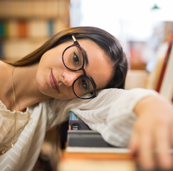 Frau mit Brille stützt ihren Kopf auf Bücher in einer Bibliothek