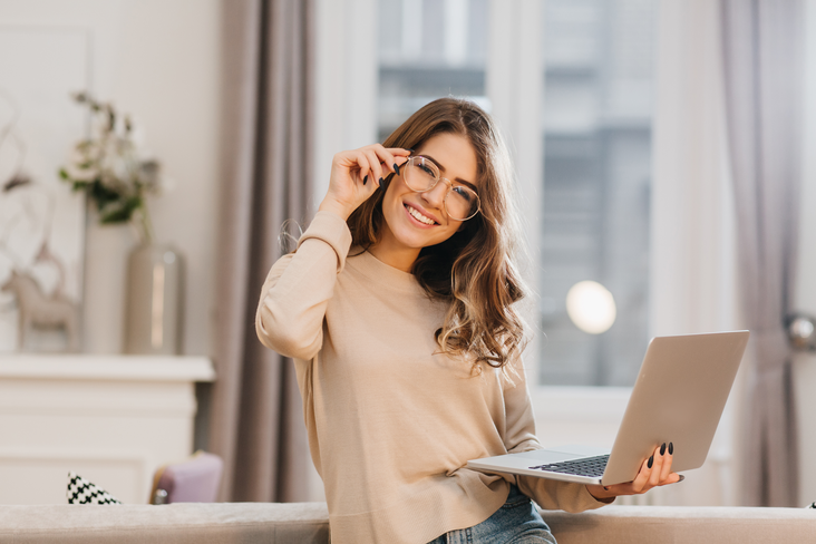 Girl with beige shirt, touching her glasses, holding laptop with smile